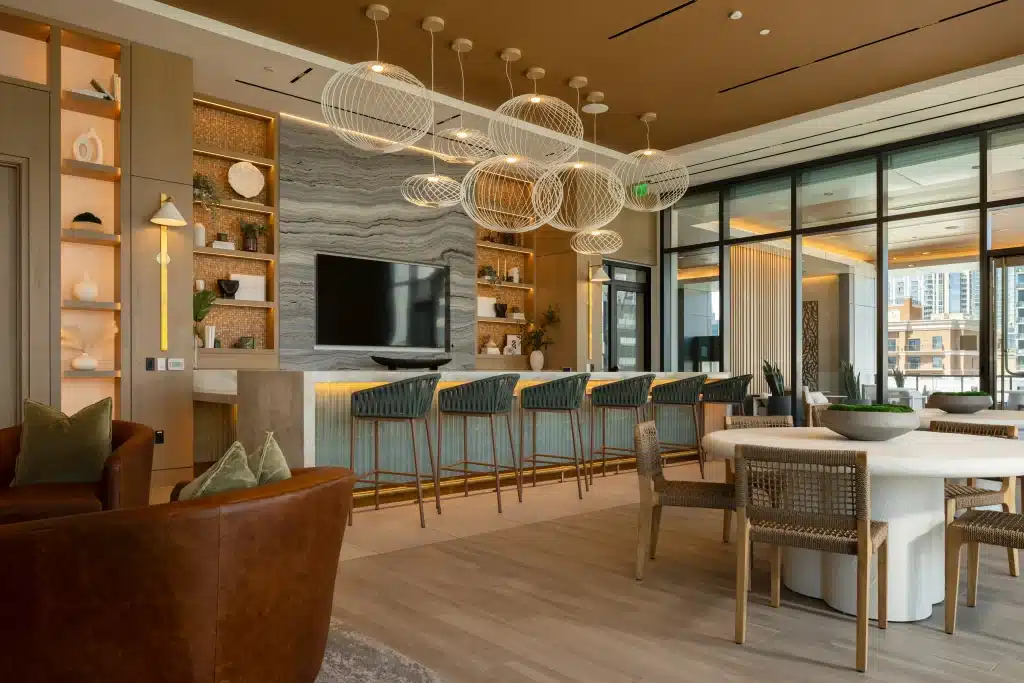 Resident lounge at 415 Colorado in Austin, with a bar counter and stools, globe-shaped wire pendant lights, a veined feature wall with lit shelving and a round dining table