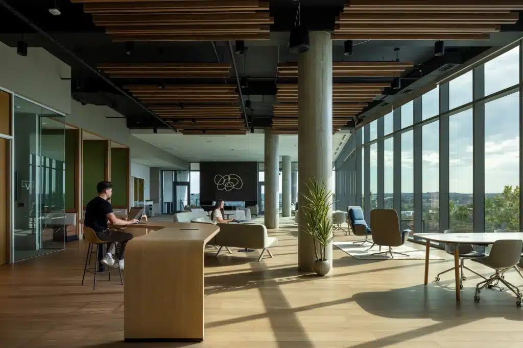 Open coworking space at The Travis ATX in Austin, with wood-slat ceiling baffles, concrete columns, a curved wood work counter and floor-to-ceiling windows