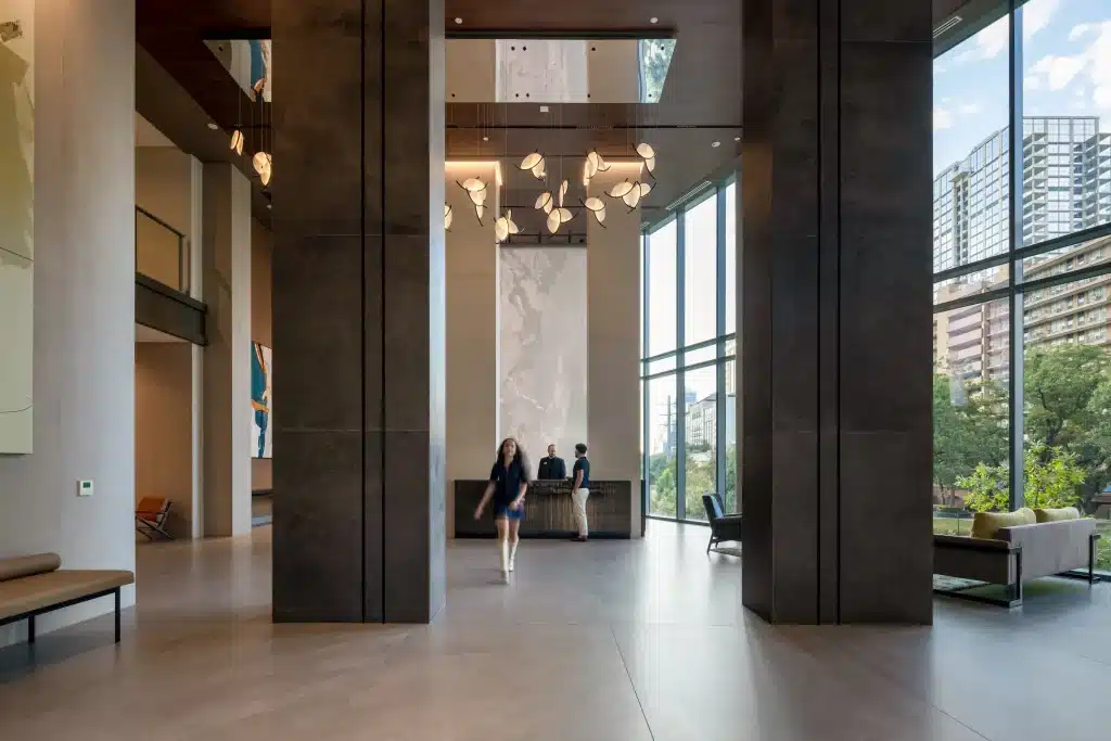 Double-height lobby and reception desk at The Travis ATX in Austin, with dark-clad columns, a cluster of leaf-shaped pendant lights and a full-height glass wall