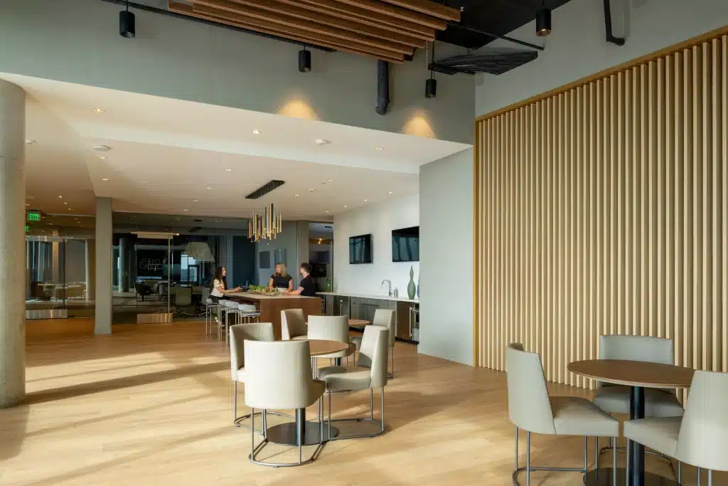 Kitchen and café seating in The Travis ATX coworking space, Austin, with round tables, an island under a gold-toned linear pendant and a tall wood-slat feature wall