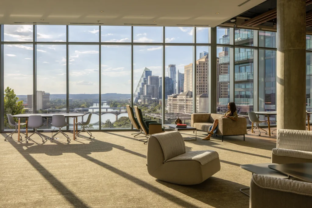 Coworking lounge at The Travis ATX with floor-to-ceiling windows framing the downtown Austin skyline and Lady Bird Lake, furnished with lounge chairs and work tables